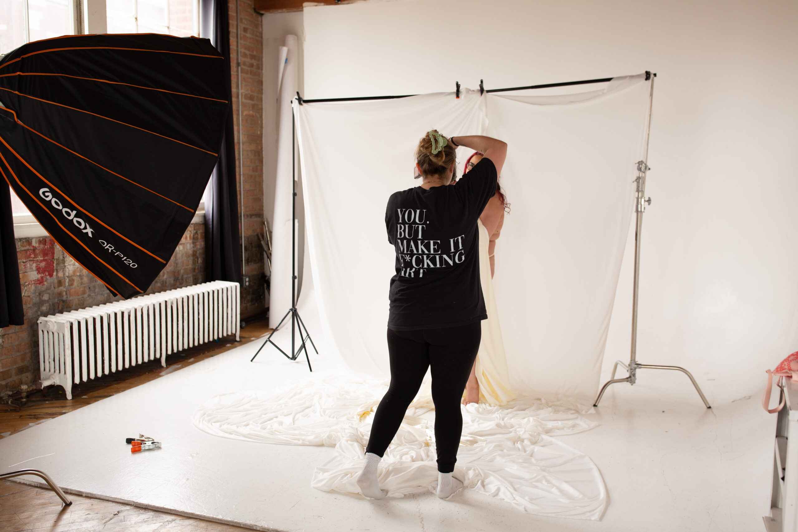 Bri Valiente Boudoir photographer photographing client in her Chicago studio with softbox lighting and white fabric backdrop. Shirt says ‘YOU. BUT MAKE IT F*CKING ART.’